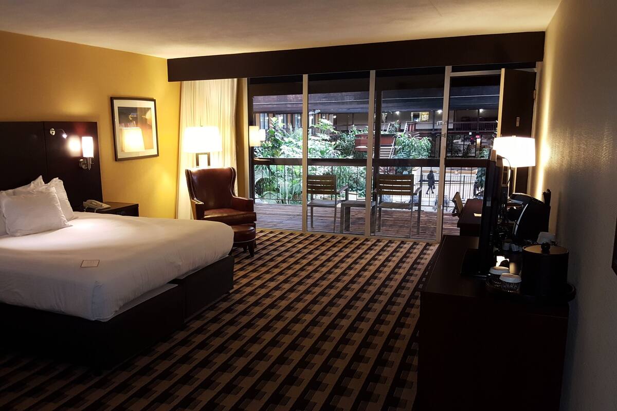 Habitación de hotel con una cama grande, una silla, un escritorio con lámpara y vistas a un balcón con mobiliario exterior.