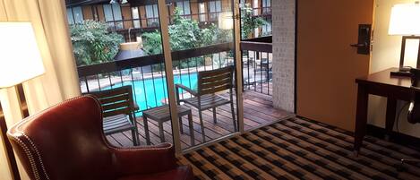 A hotel room with a view of an outdoor swimming pool, a leather chair, a small table, and a patterned carpet.