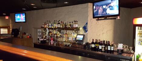 Una zona de bar con varias estanterías llenas de botellas de licor, una caja registradora y un televisor montado en la pared.