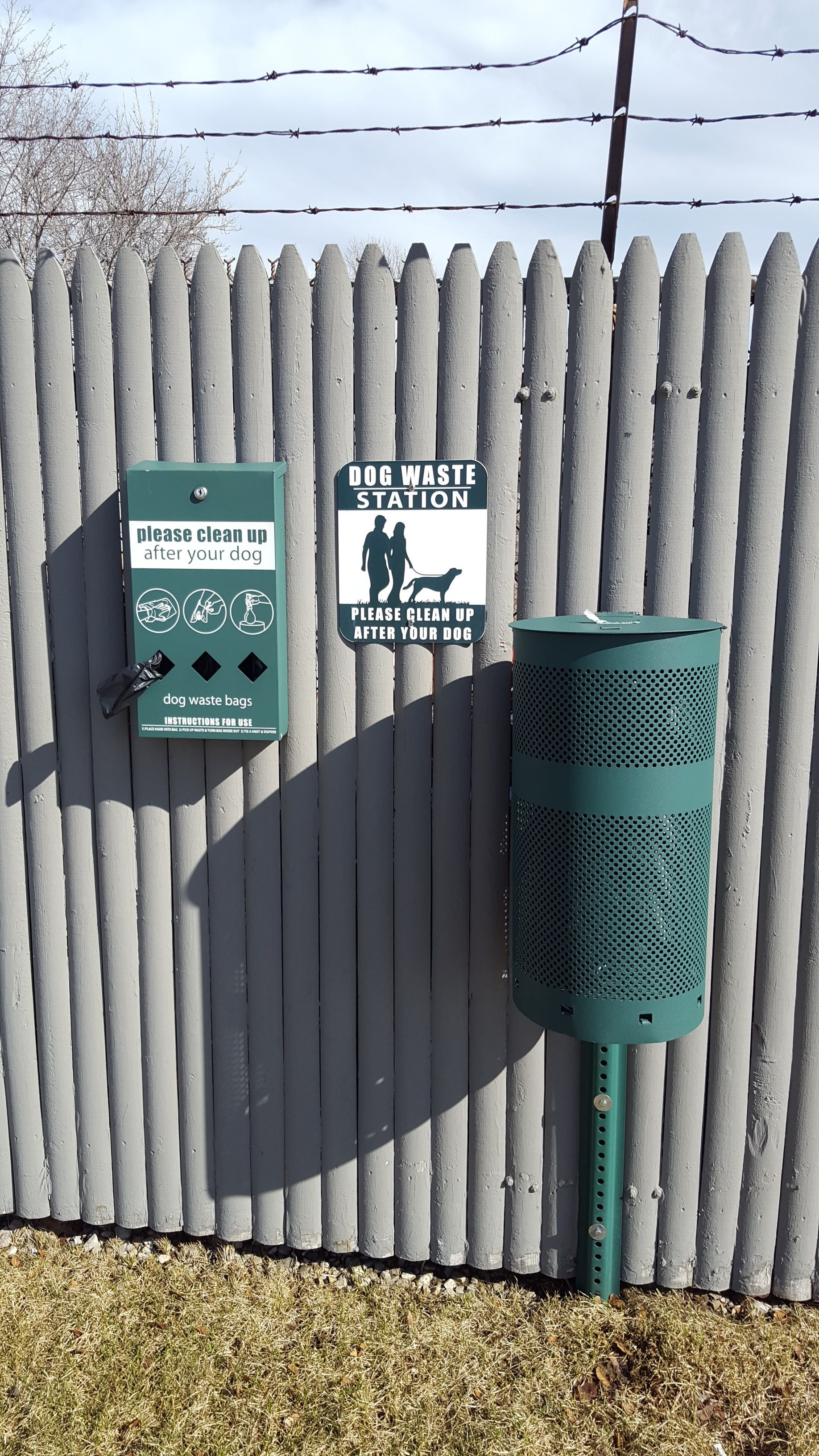 A dog waste station with a sign instructing to clean up after your dog, a trash can, and a fence.