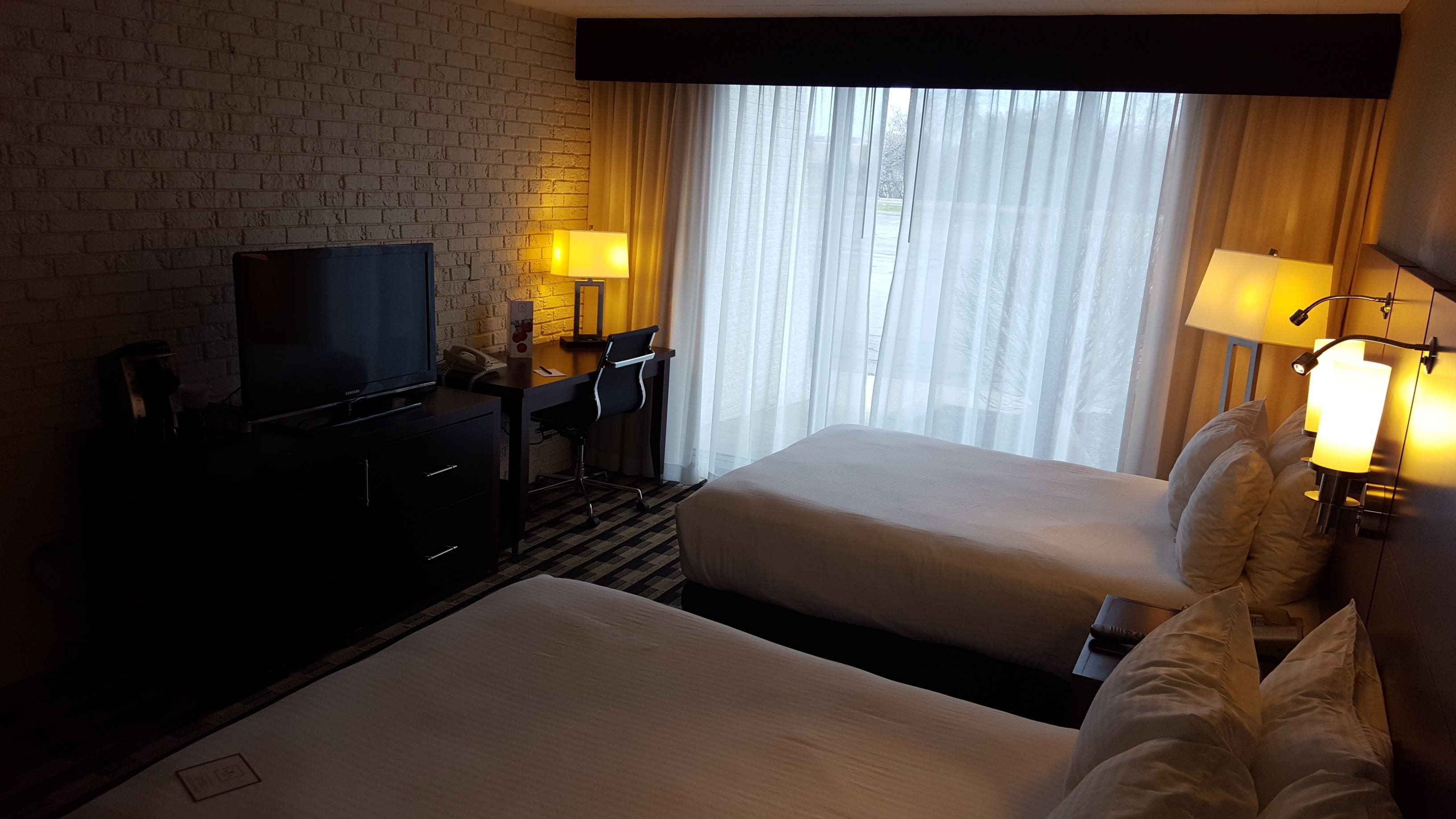 A hotel room with two beds, a TV, a desk with a chair, and a window with curtains.