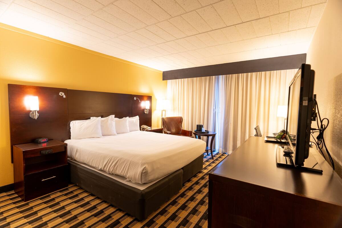Una habitación de hotel con una cama grande, un escritorio con computadora, una silla, una mesita de noche y una lámpara.