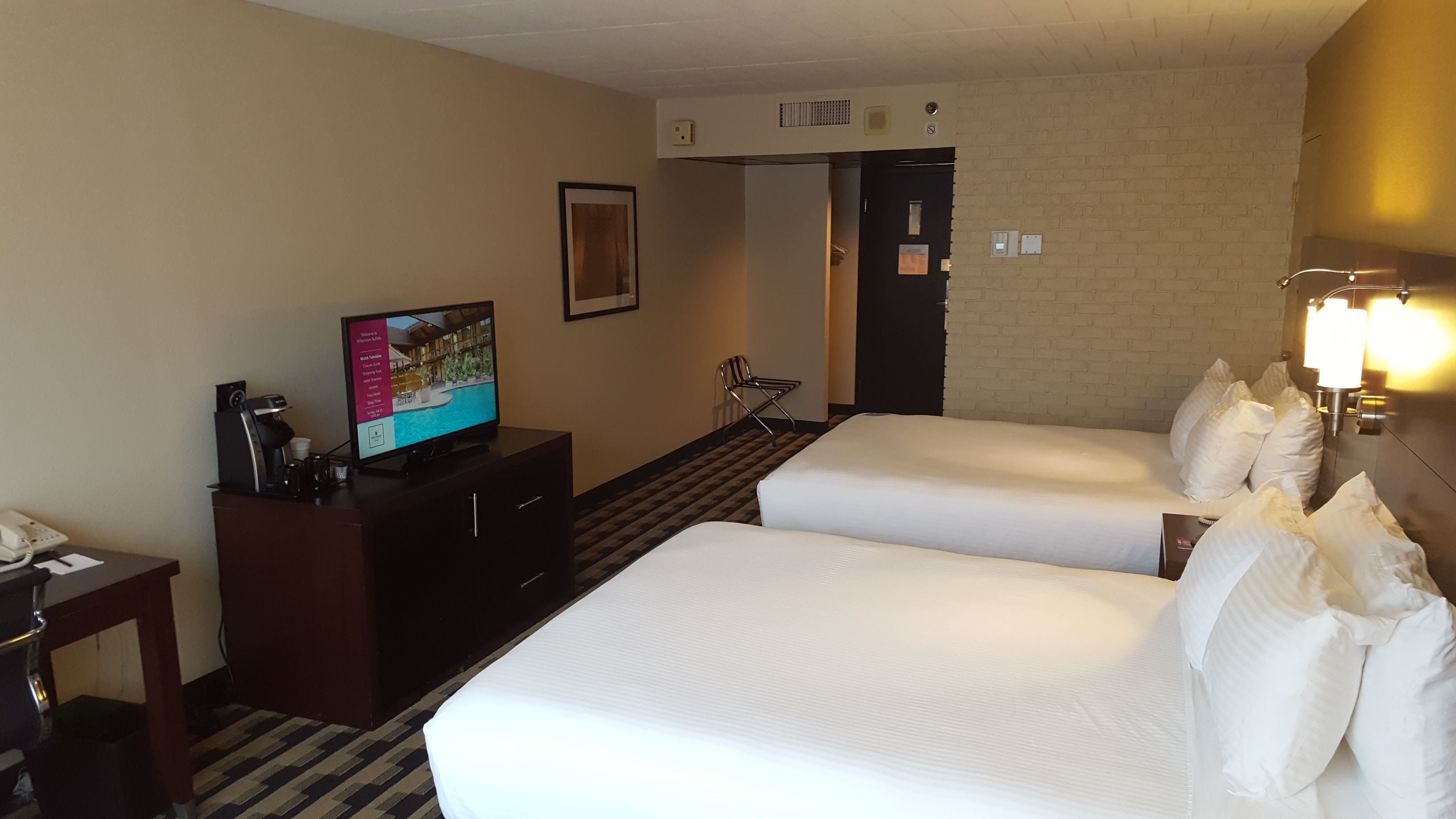 Une chambre d’hôtel avec deux lits, un bureau avec une machine à café, un téléviseur diffusant une brochure de l’hôtel et un mur décoré d’un motif en briques.