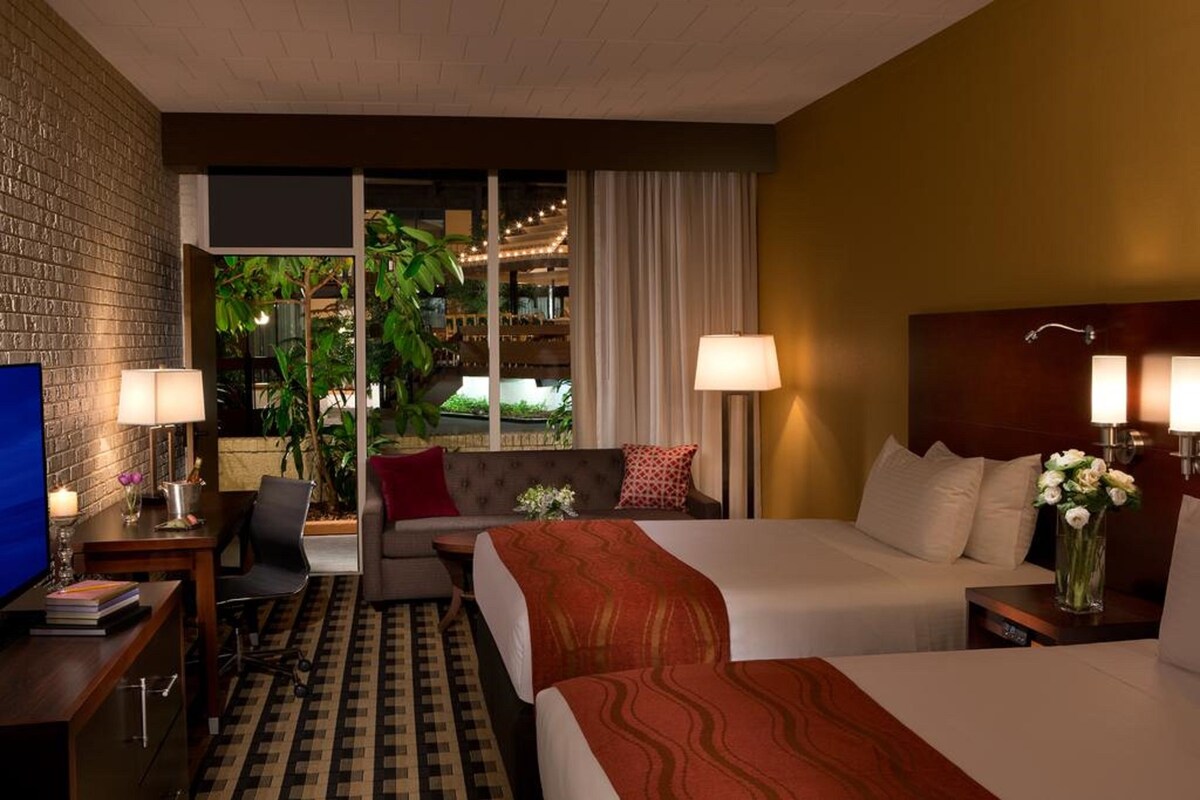 A hotel room with two beds, a desk, a TV, and a view of the outdoor area.