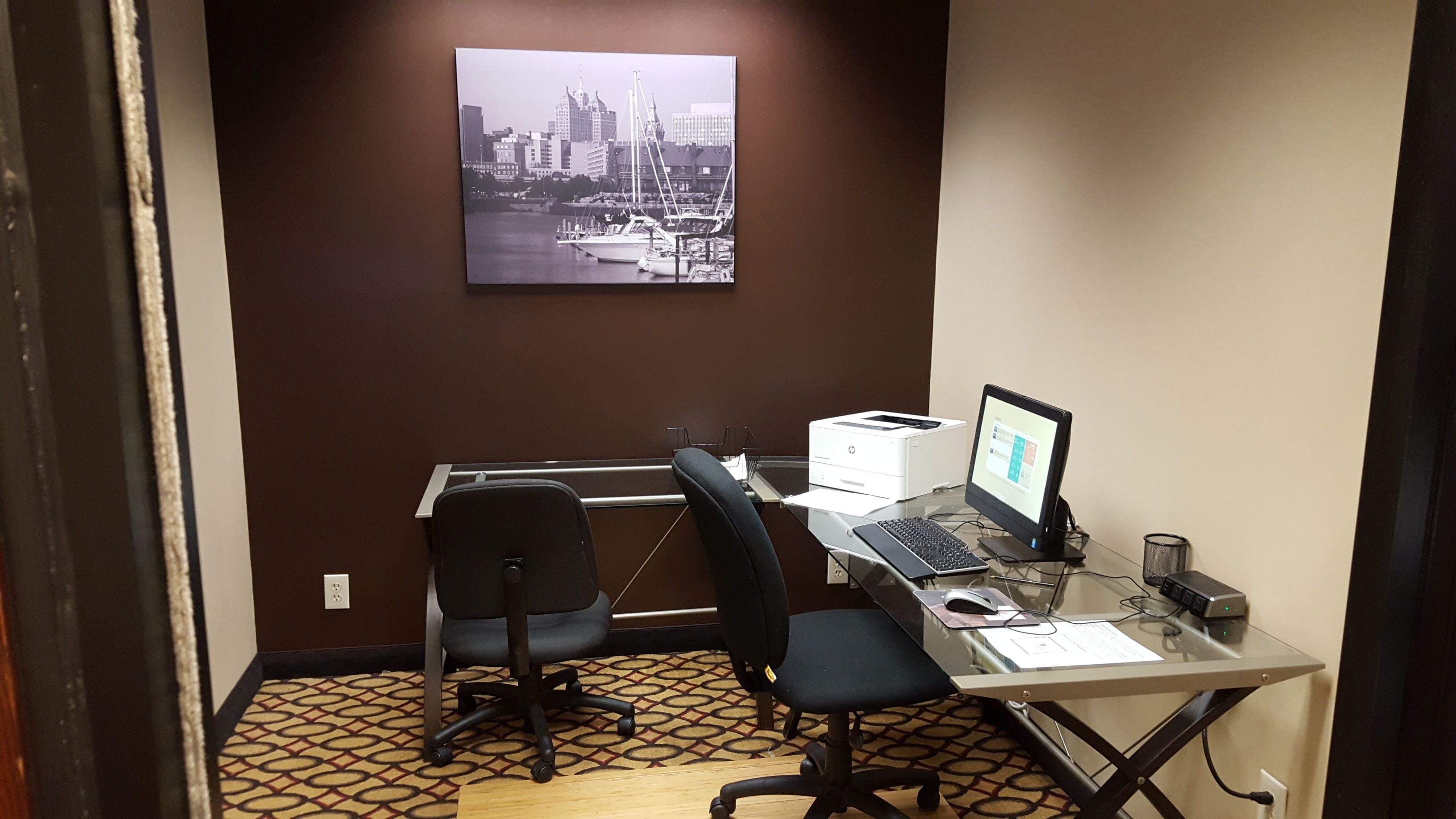 Un pequeño espacio de oficina con un escritorio, dos sillas, una computadora e impresora. Una fotografía de un paisaje urbano cuelga en la pared.