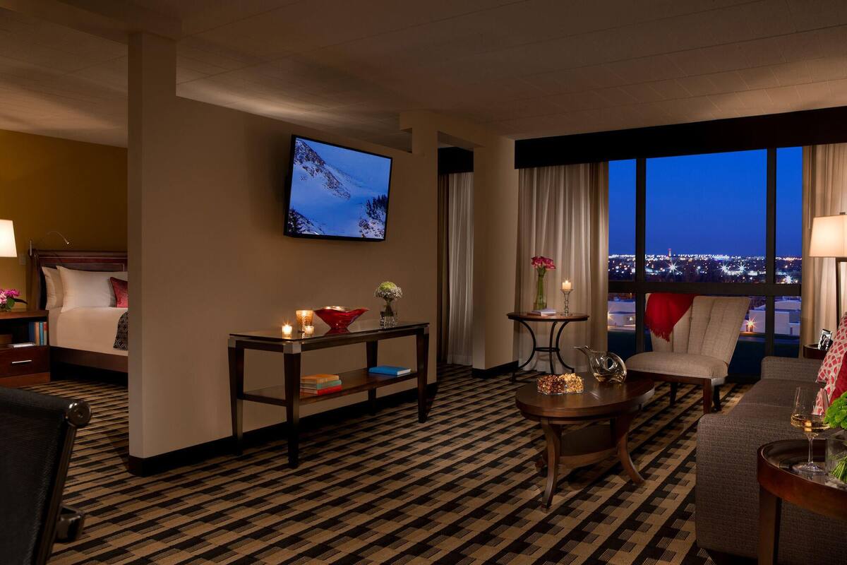 Habitación de hotel con cama, televisor montado en la pared, mesita con un florero y velas, una silla y vista a la ciudad de noche.