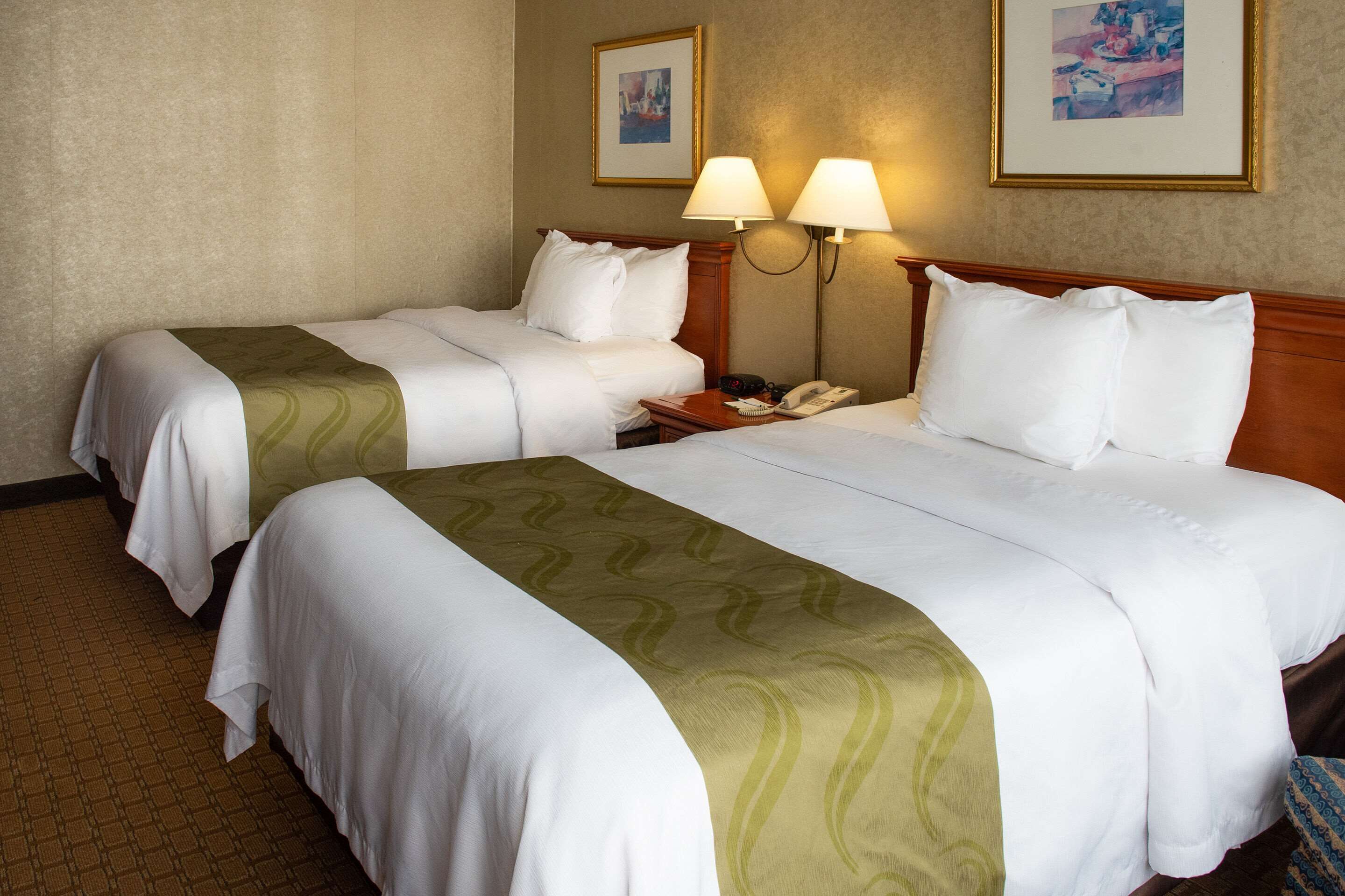 A hotel room with two beds, each with white linens and a green accent. There are two wall-mounted lamps, a telephone, and framed artwork on the walls.