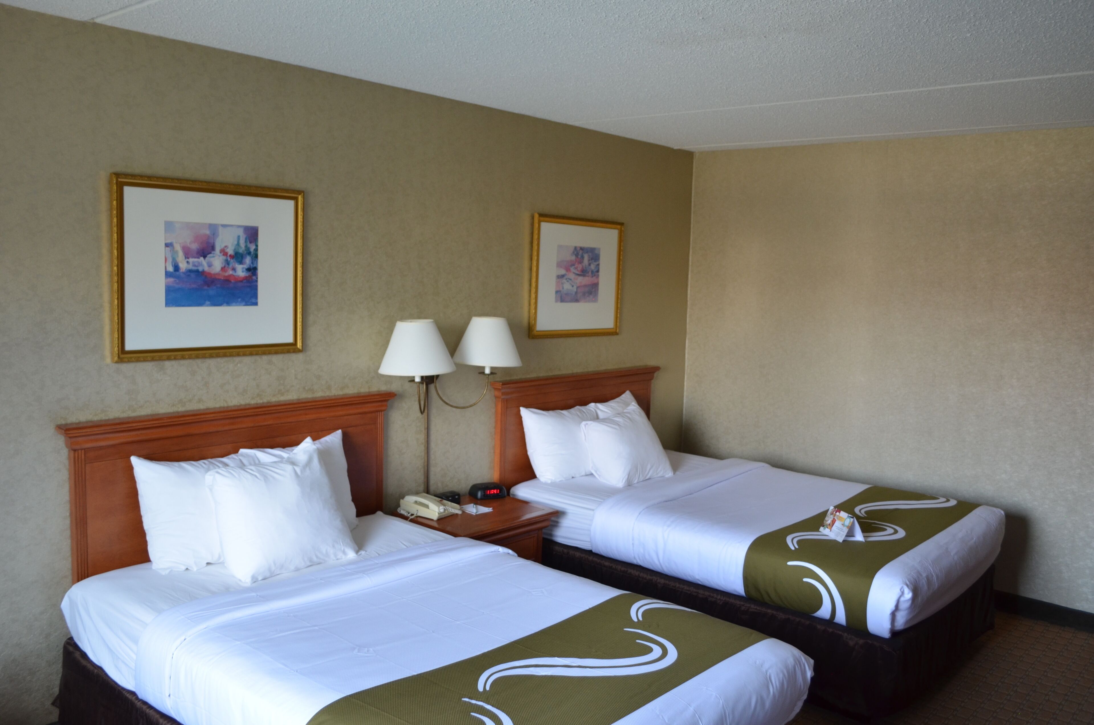 Quarto de hotel com duas camas, uma cabeceira de madeira, mesa de cabeceira com abajur e duas fotos emolduradas na parede.