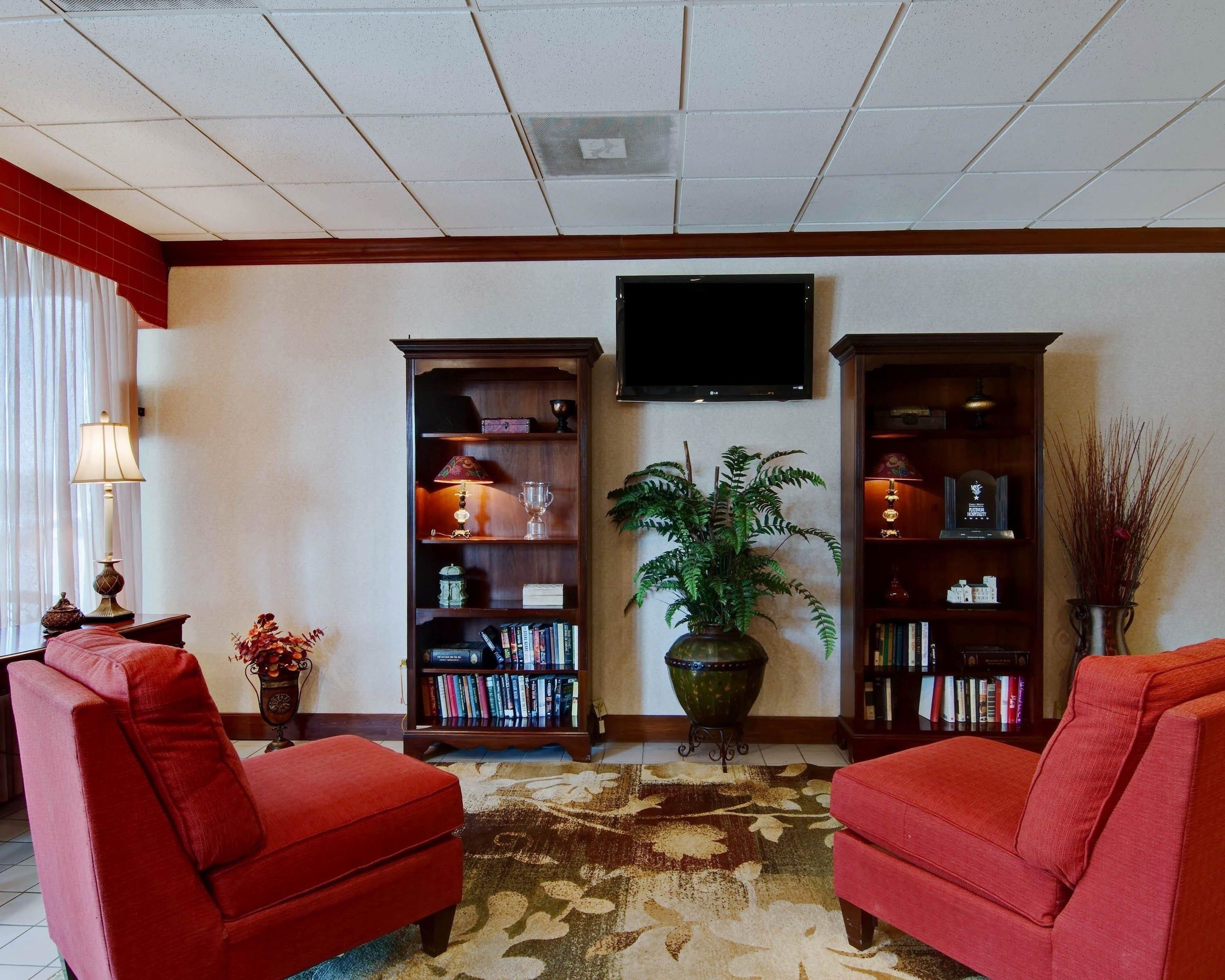 A hotel lobby with two red armchairs, a large potted plant, and two tall bookshelves filled with books and decorative items.