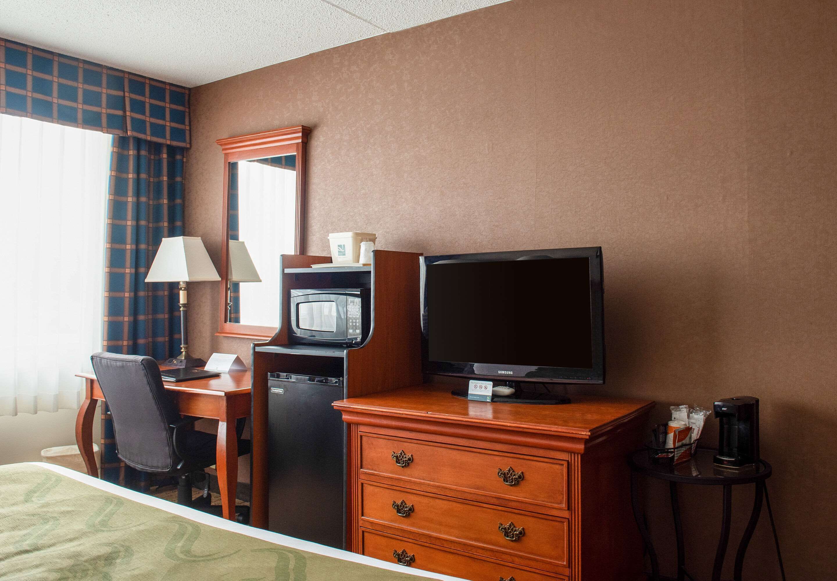 A hotel room with a bed, a desk with a computer, a television, a microwave, a coffee maker, and a dresser with drawers.