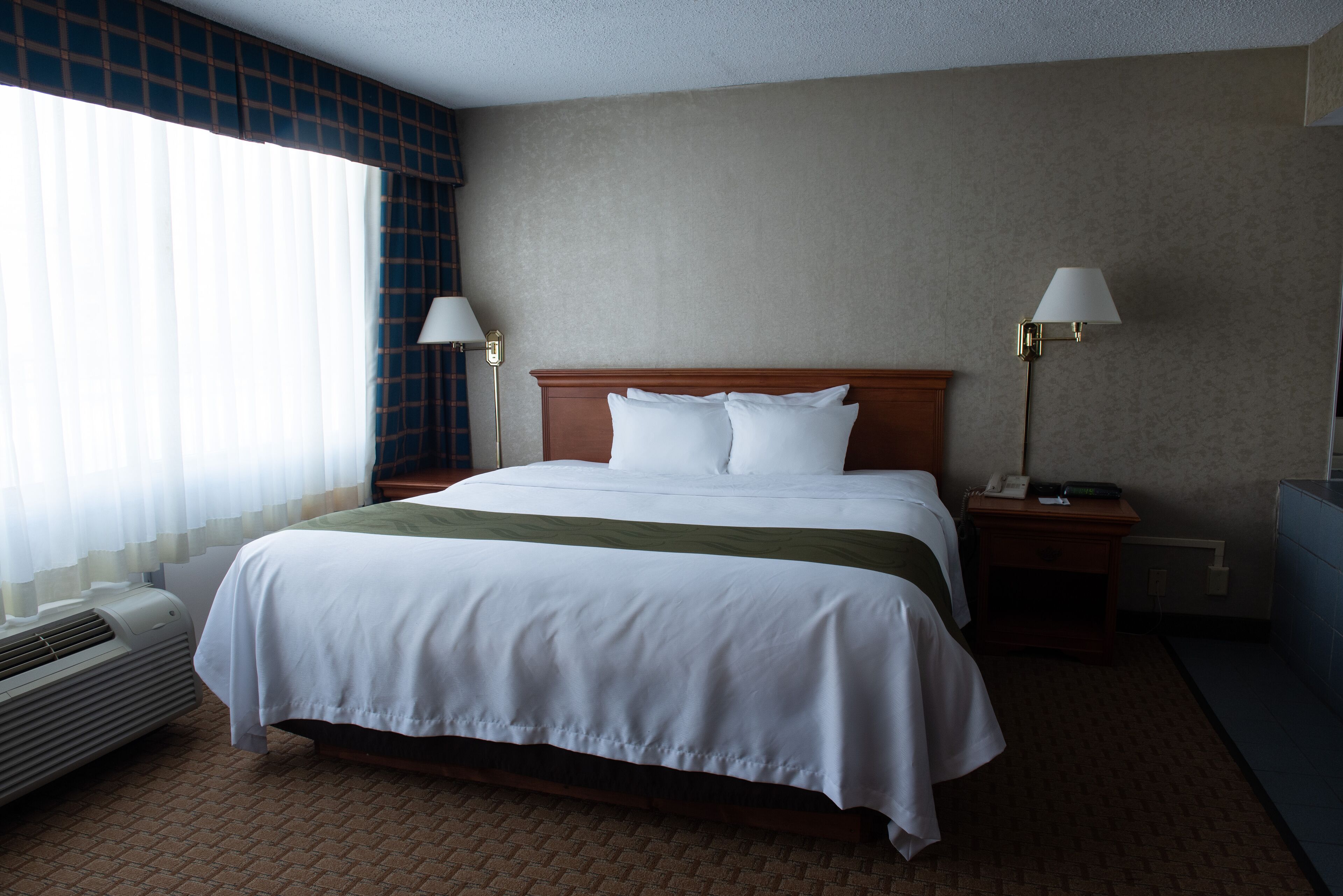 A hotel room with a large bed, two bedside tables with lamps, a window with blinds, and a wall-mounted air conditioner.