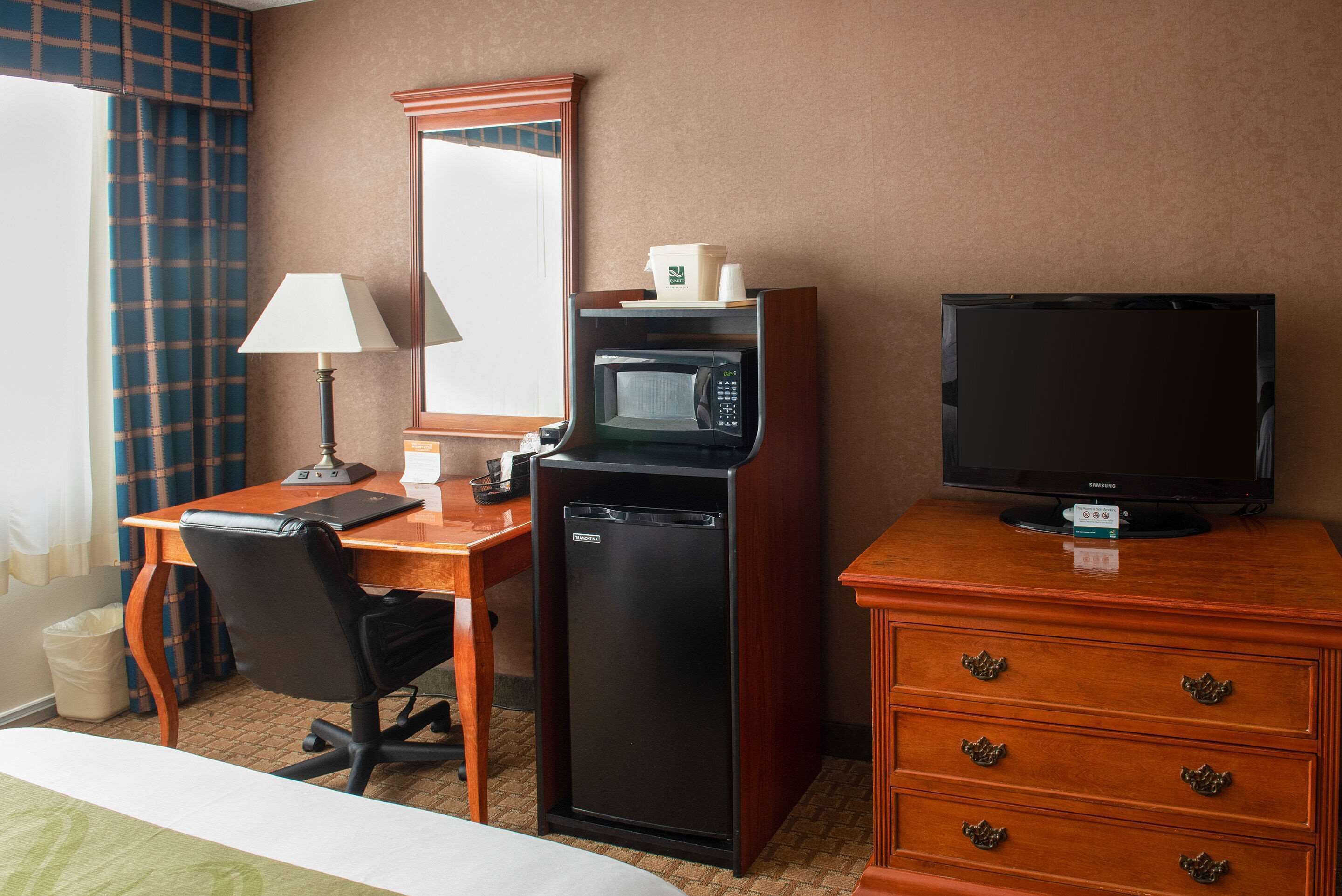 A hotel room with a desk, a microwave, a television, a dresser, a chair, and a lamp.