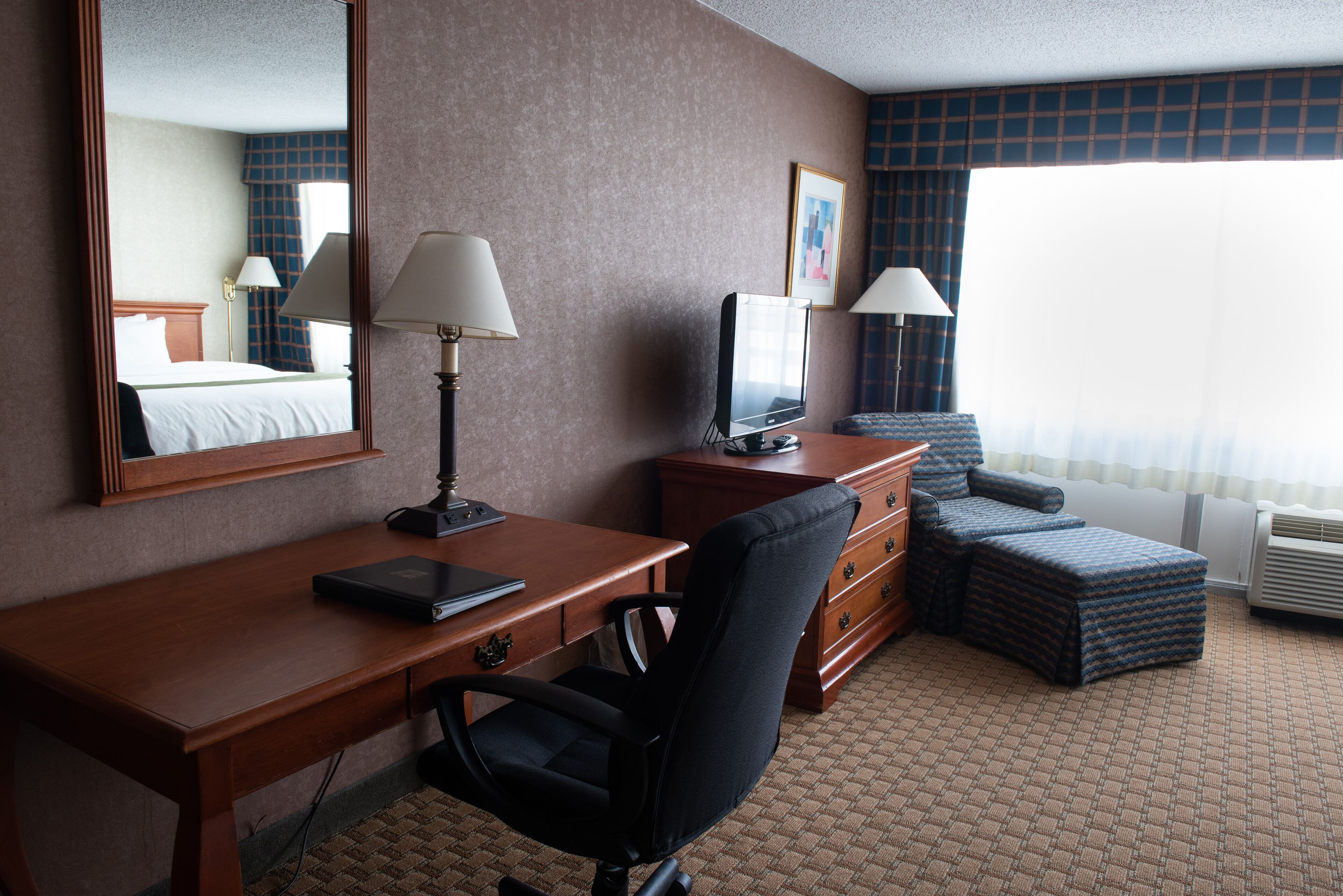 Une chambre d’hôtel avec un bureau en bois, un fauteuil de bureau noir, une télévision, une commode et une fenêtre avec des rideaux.