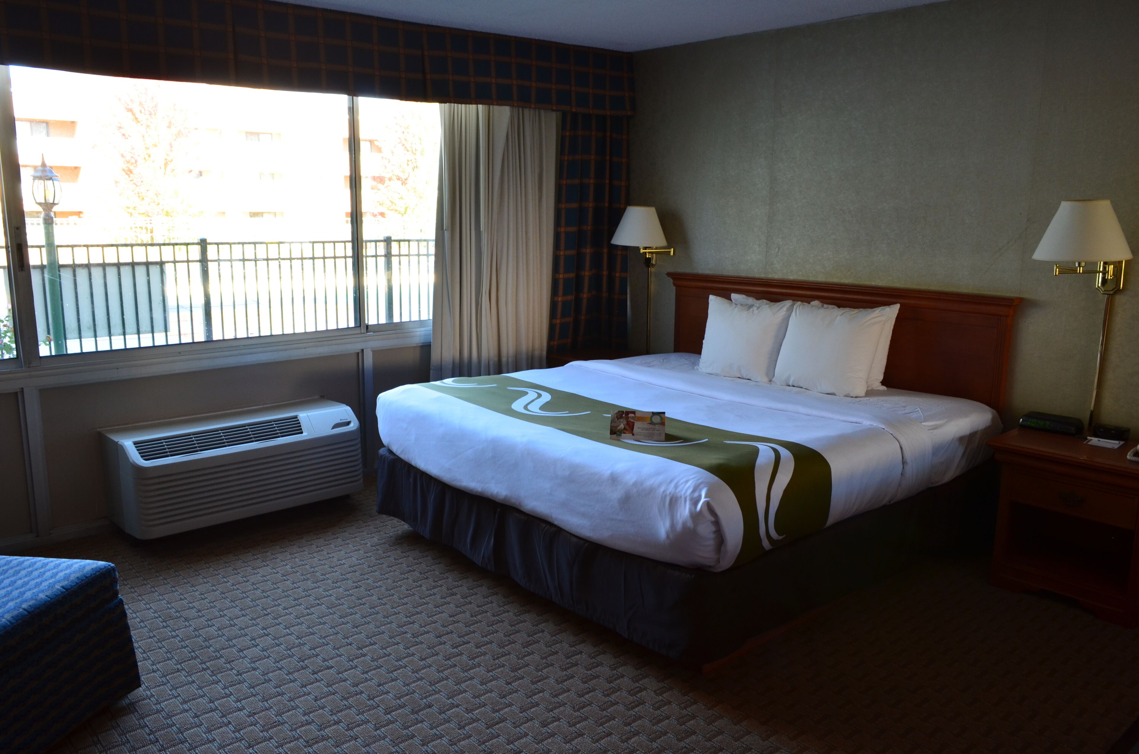 A hotel room with a large bed, two bedside lamps, a window with curtains, and a wall-mounted air conditioner.