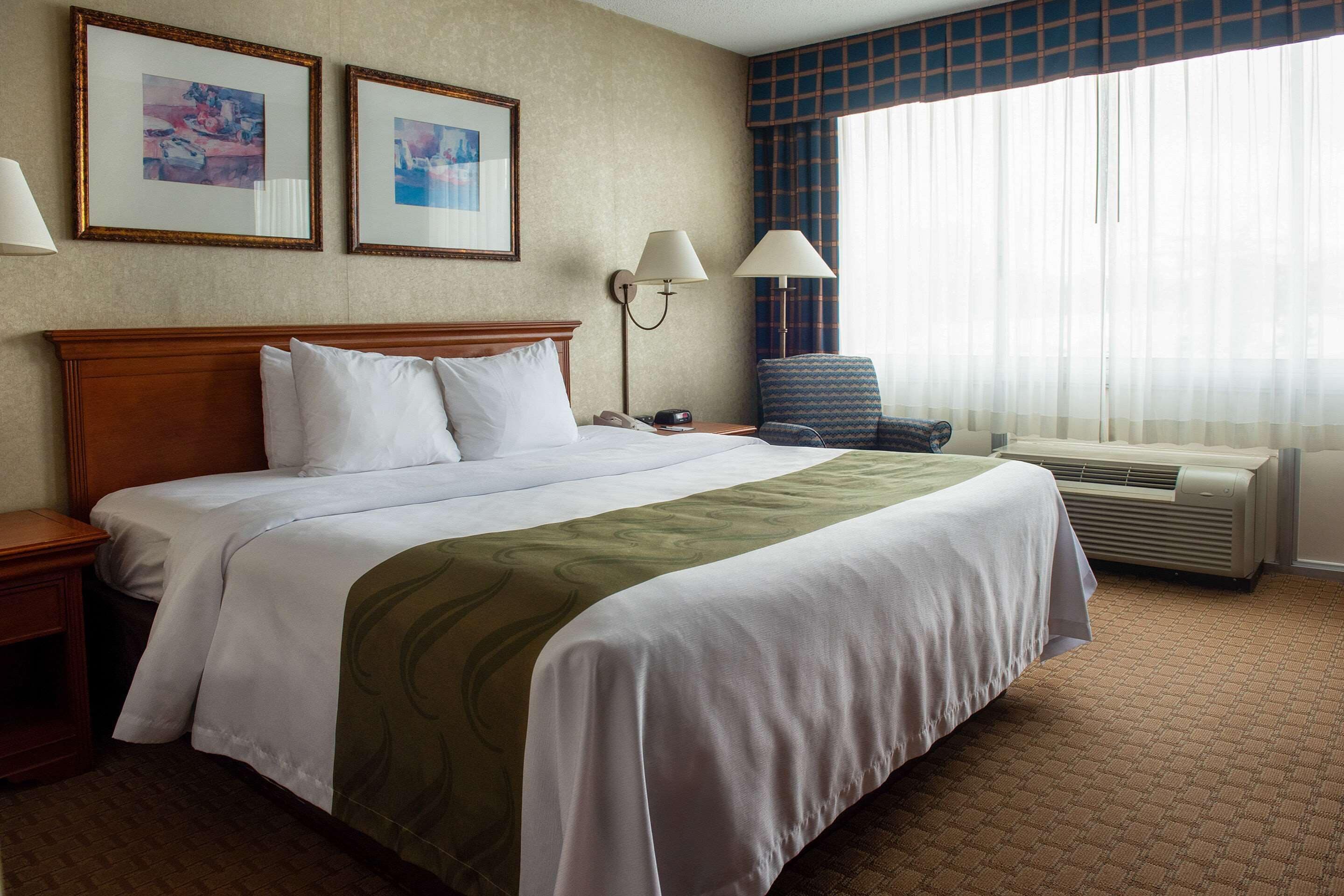 A hotel room with a large bed, two framed pictures on the wall, a nightstand, a chair, and a window with curtains.