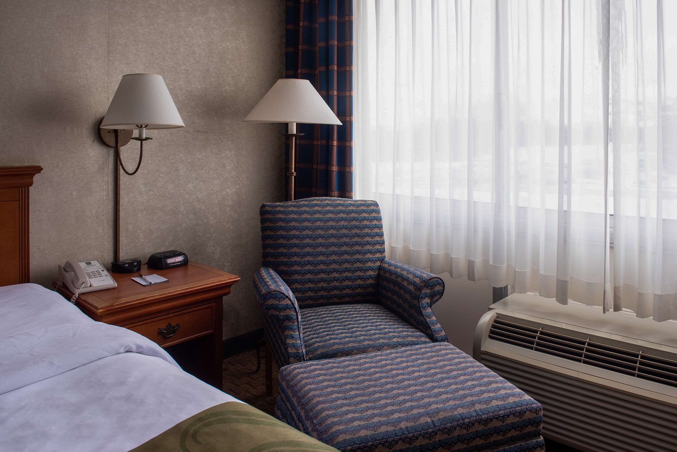 A hotel room with a bed, a desk with a lamp, a chair, and an air conditioning unit.