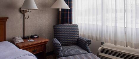 A hotel room with a bed, a desk with a lamp, a chair, and an air conditioning unit.