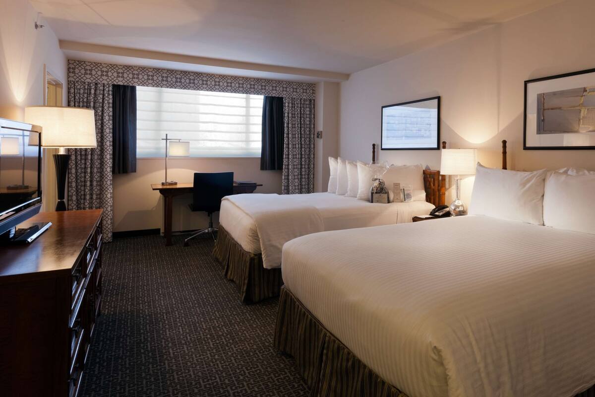 A hotel room with two beds, a desk, a chair, a TV, and a lamp.