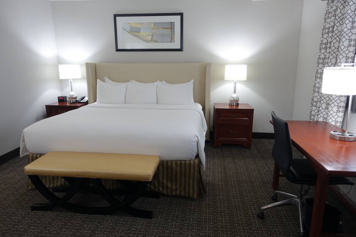A hotel room with a large bed, bedside tables with lamps, a desk with a chair, and a bench.