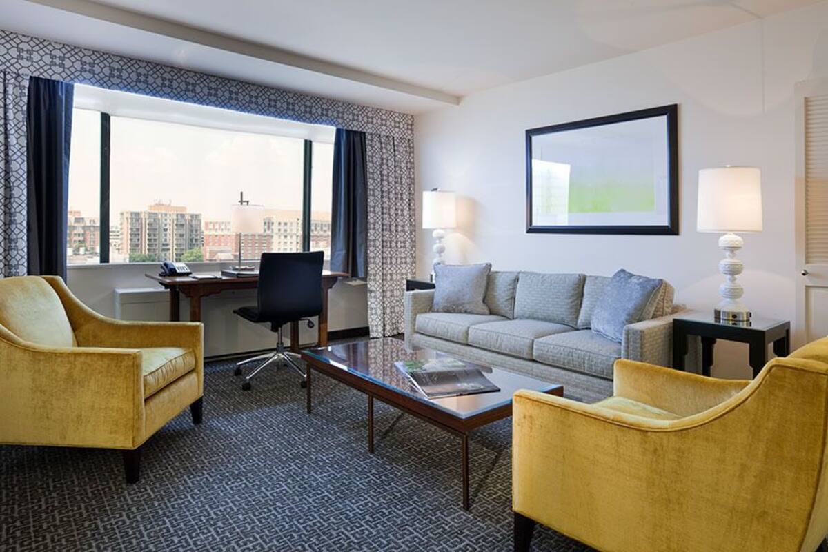 A hotel room with a sofa, two armchairs, a coffee table, a desk, and a lamp.