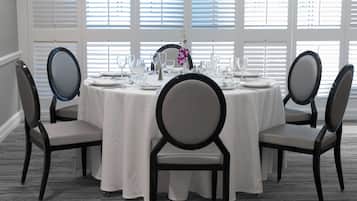 A round table set for six with white tablecloths, six chairs with black frames and grey cushions, and a vase with purple flowers.