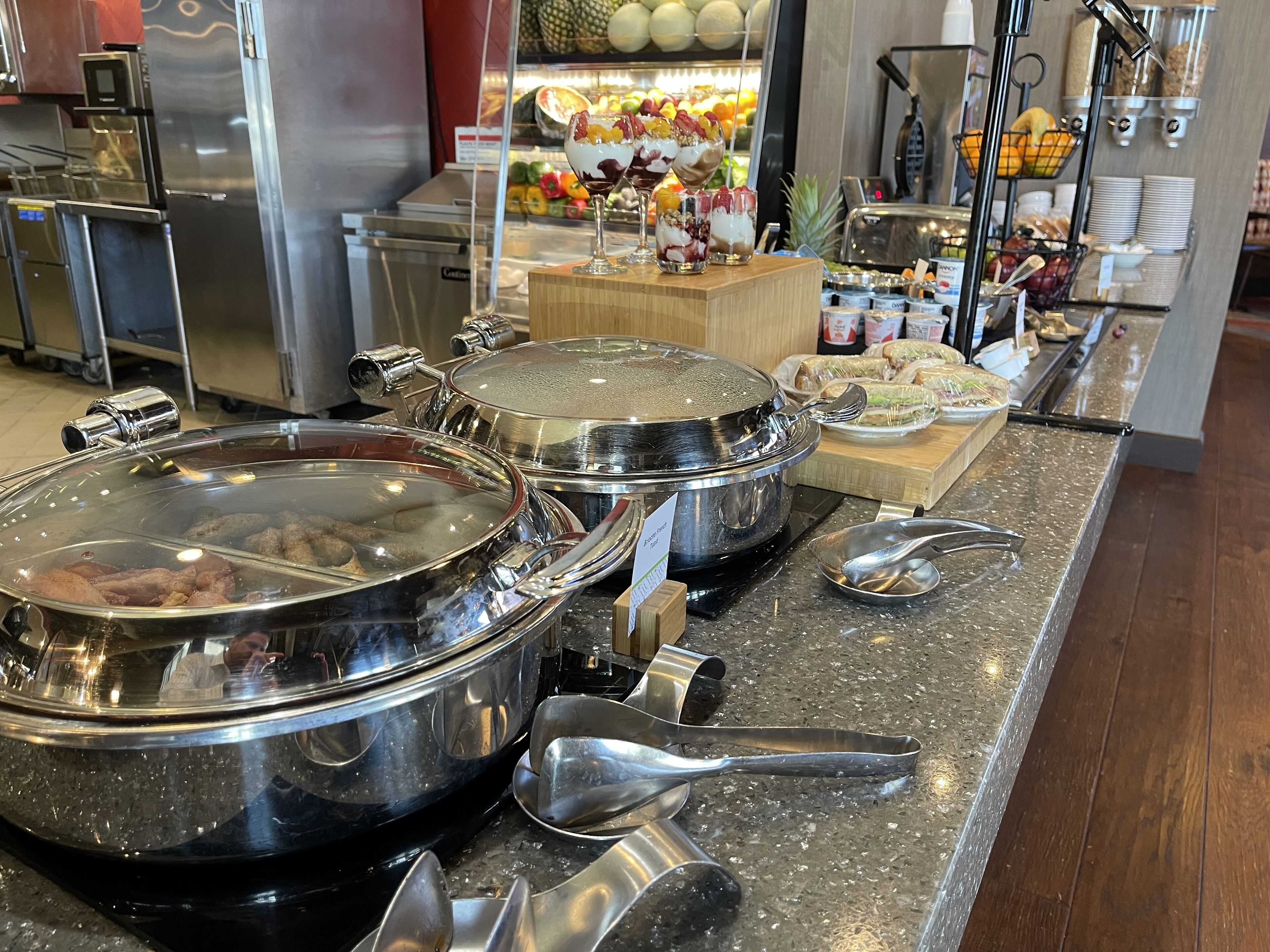 A buffet setup with covered food containers, serving utensils, and a fruit display in the background.