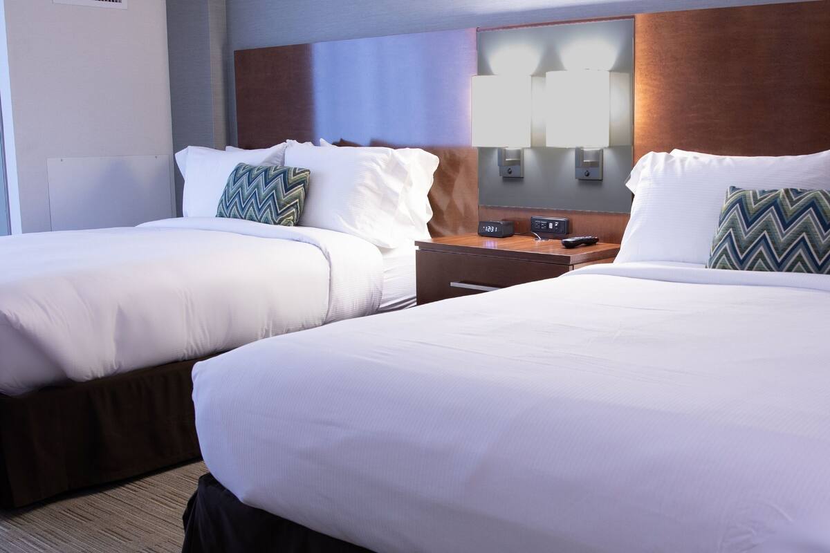 Hotelkamer met twee bedden, elk voorzien van witte lakens en decoratieve kussens. Een houten hoofdbord, een nachtkastje met een klok en een spiegel boven het hoofdbord.
