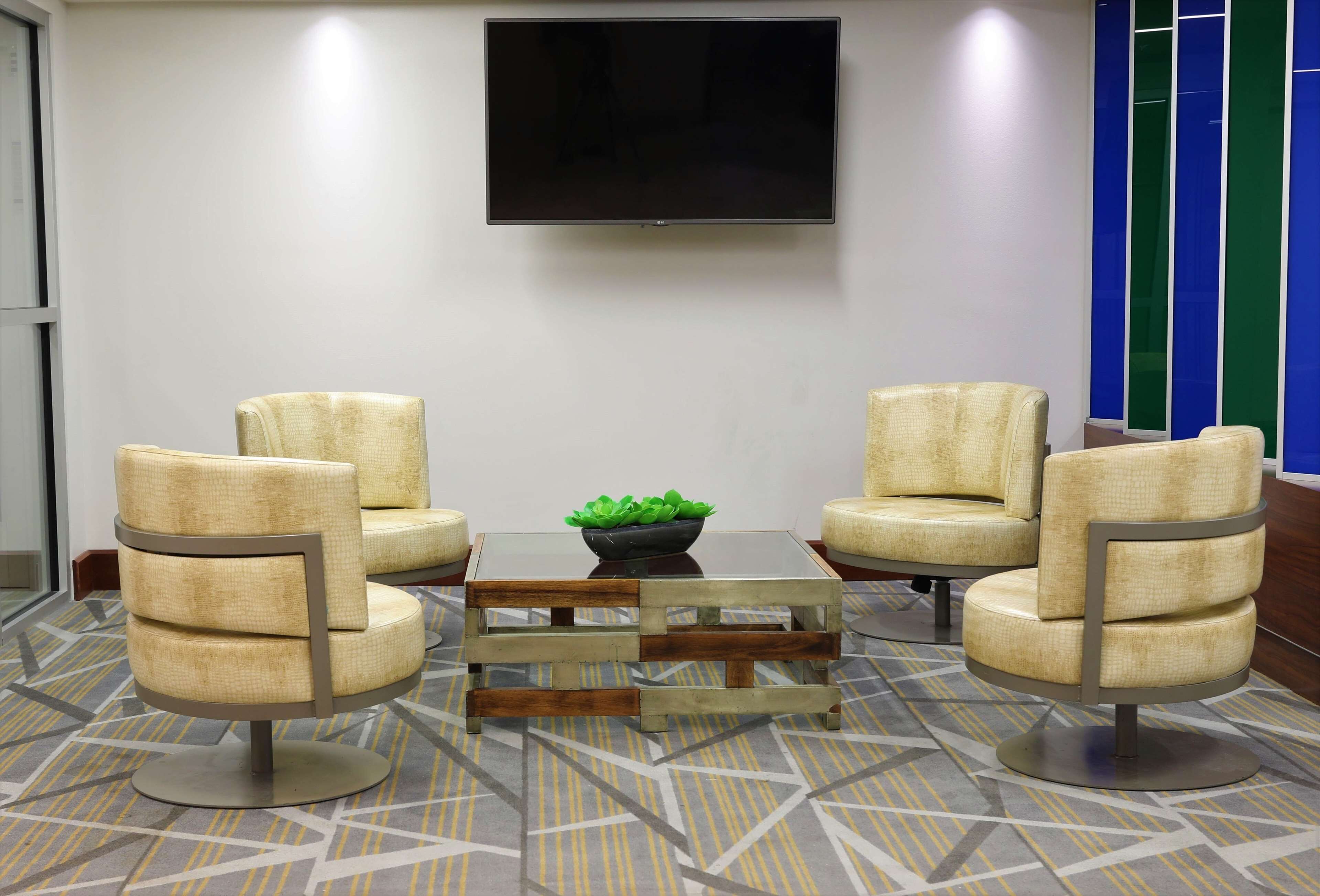 Un área de espera moderna con cuatro sillones color beige, una mesa de centro de vidrio con marco de madera y un televisor de pantalla plana montado en la pared.