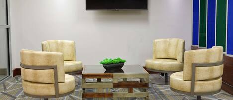 Un área de espera moderna con cuatro sillones color beige, una mesa de centro de vidrio con marco de madera y un televisor de pantalla plana montado en la pared.
