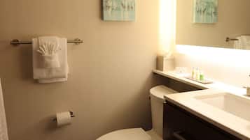 Un baño con inodoro, toallero con toalla doblada, dispensador de papel higiénico, lavabo con artículos de aseo y dos cuadros colgados en la pared.