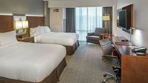 Een hotelkamer met twee bedden, een bureau met stoel, een televisie en een groot raam met gordijnen.