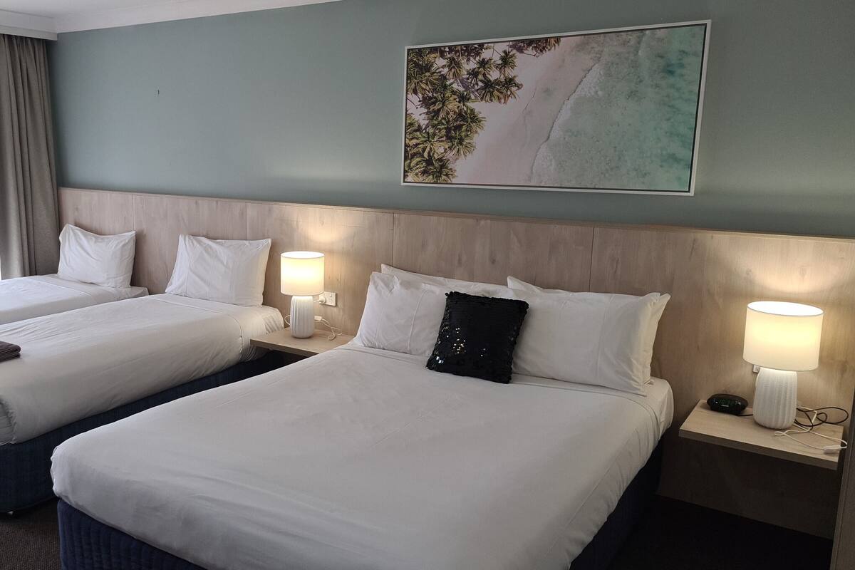 A hotel room with two beds, a wooden headboard, a painting of a beach, and a window with curtains.