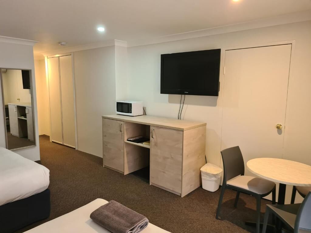 A hotel room with a bed, a desk with a microwave, a TV, a small table with chairs, and a trash bin.
