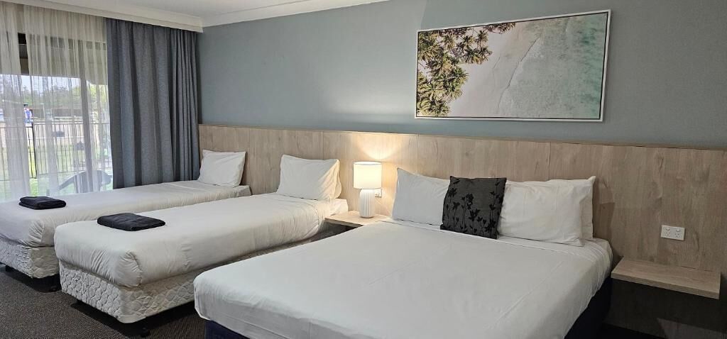 A hotel room with two beds, a large window with curtains, a wall-mounted artwork, and a bedside table with a lamp.