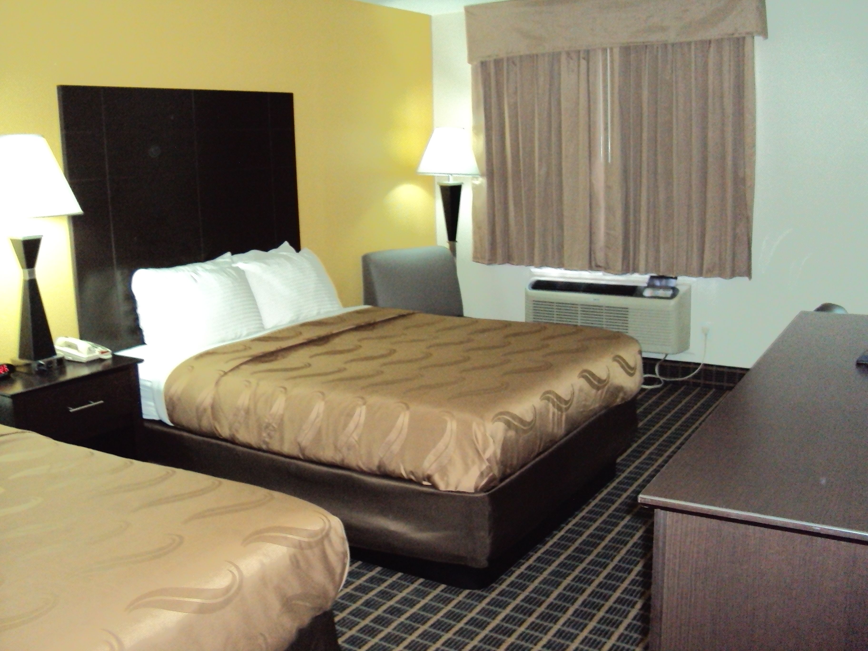 A hotel room with a large bed, a desk, a chair, a lamp, and an air conditioning unit.