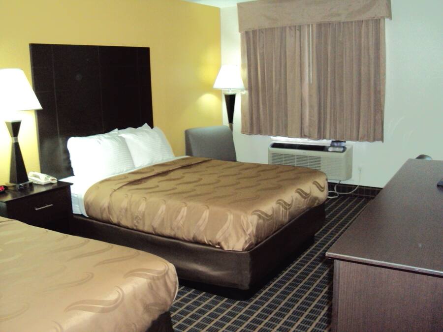 A hotel room with a large bed, a desk, a chair, a lamp, and an air conditioning unit.