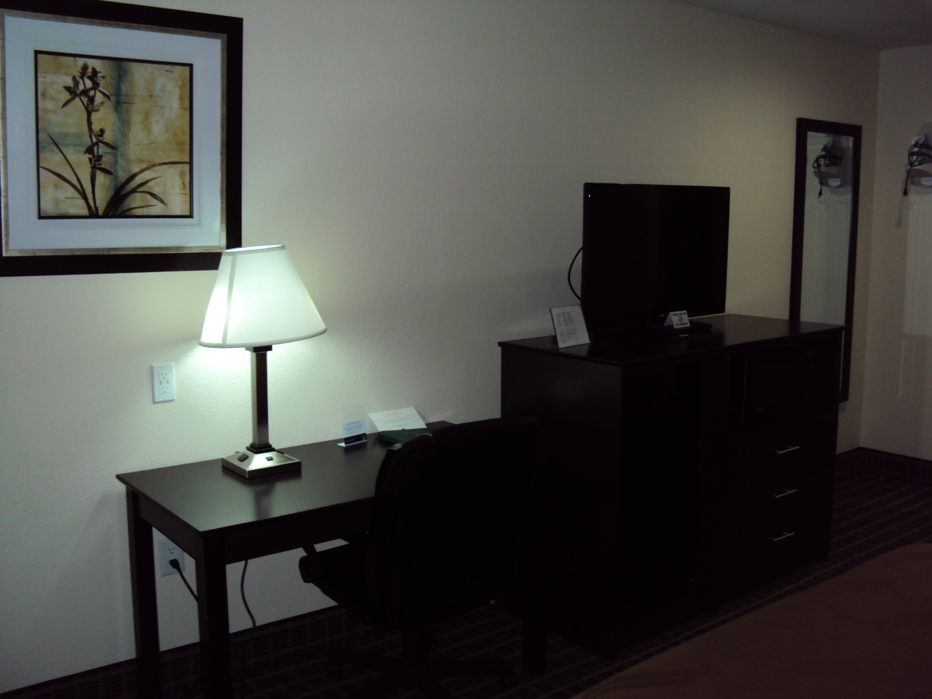 A hotel room with a desk, a chair, a lamp, a television, and a dresser.