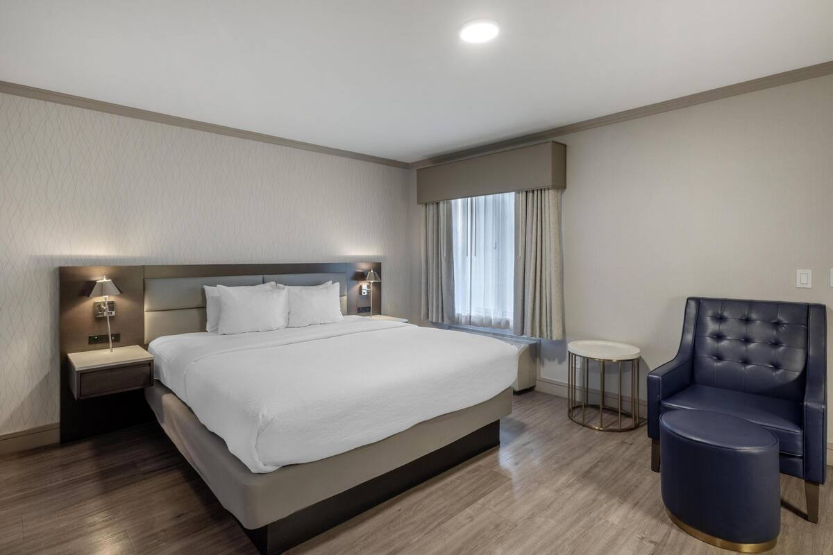 Camera d'albergo moderna con un letto grande, un tavolino con una lampada, un tavolino rotondo e una poltrona blu scuro.