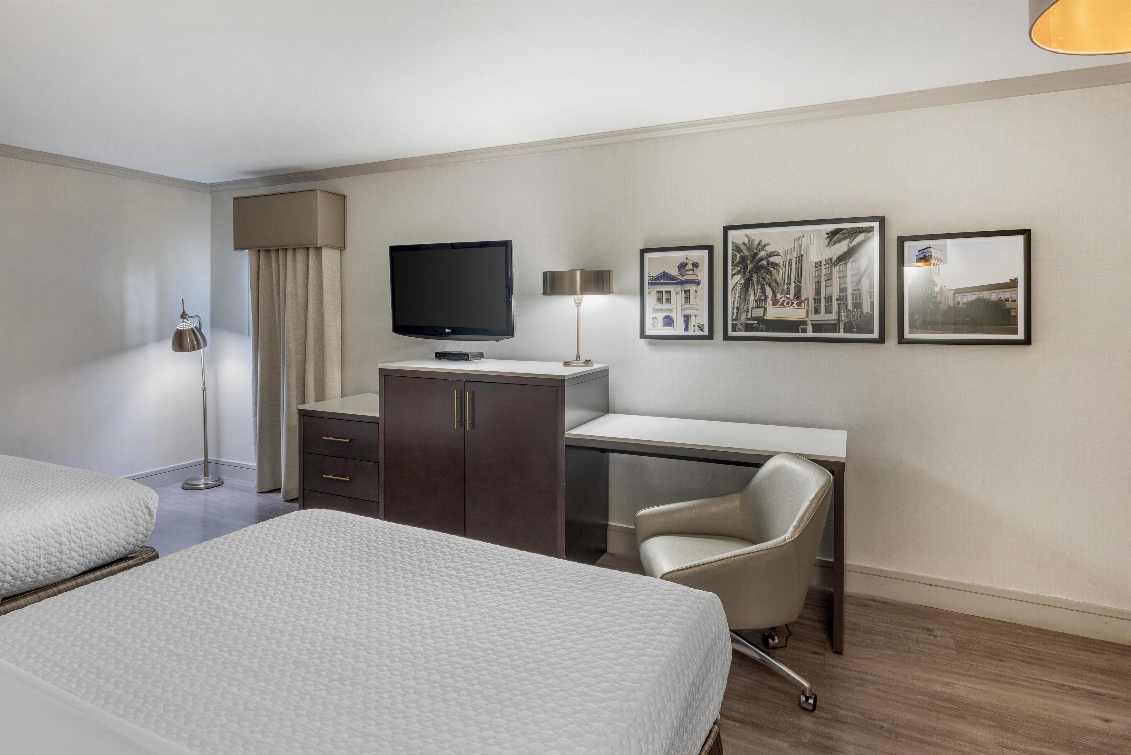 A hotel room with a bed, a desk, a chair, a TV, a lamp, and framed pictures on the wall.