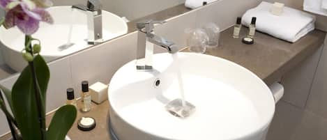Un baño con un lavamanos blanco, dos llaves, un espejo y artículos de aseo en la encimera.