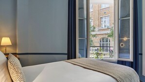 Habitació d'hotel amb un llit, una làmpada i vista a un edifici per una finestra oberta.