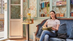 Una dona asseguda en un sofà blau a una habitació amb paper tapet floral, una prestatgeria i una taula petita amb un got de beguda.