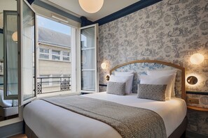 A bedroom with a large bed, patterned wallpaper, and a balcony with a view of buildings. - Hôtel de Neuve Le Marais by Happyculture (Paris)