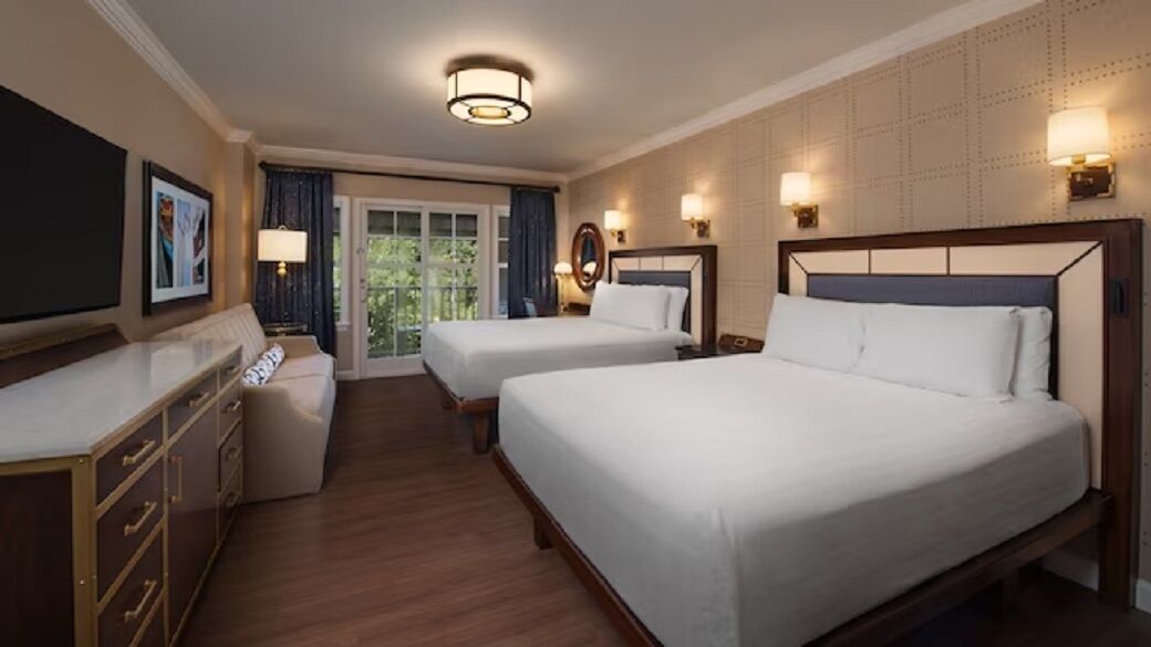 A hotel room with two beds, a desk, a dresser, and a view of the outdoors.