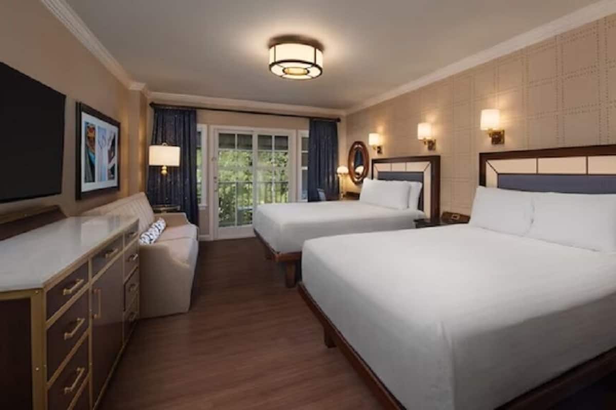 A hotel room with two beds, a desk, a dresser, and a view of the outdoors.