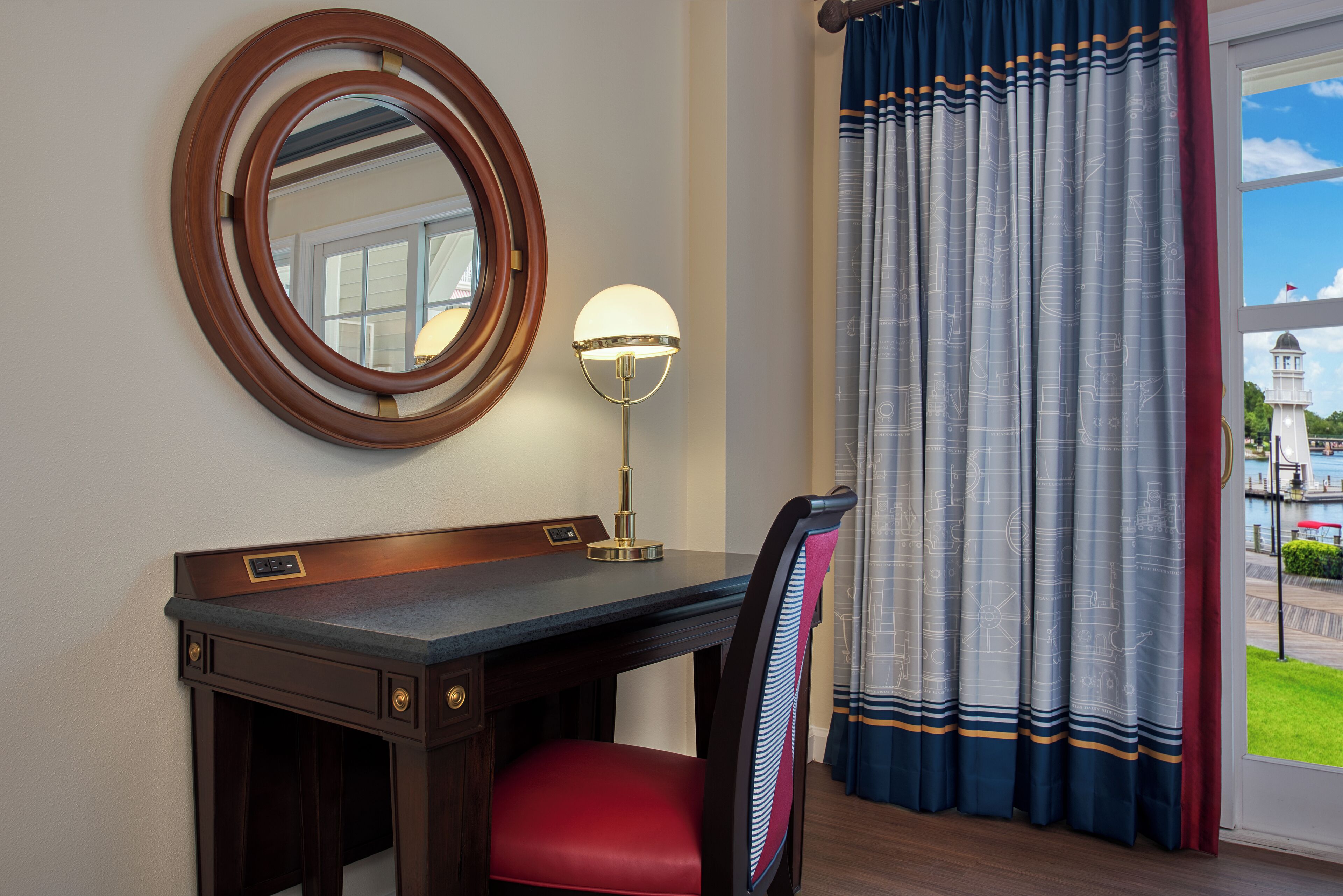 A hotel room with a desk, a chair, a round mirror, and a lamp.