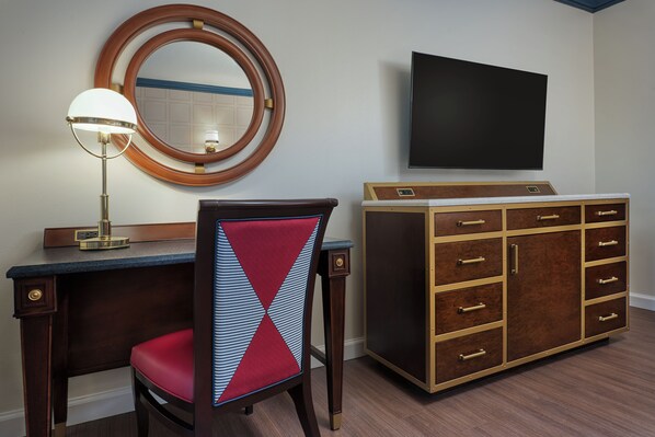 Una habitación de hotel con escritorio, una silla con cojín rojo, un aparador de madera con cajones, un espejo redondo y un televisor de pantalla plana montado en la pared.