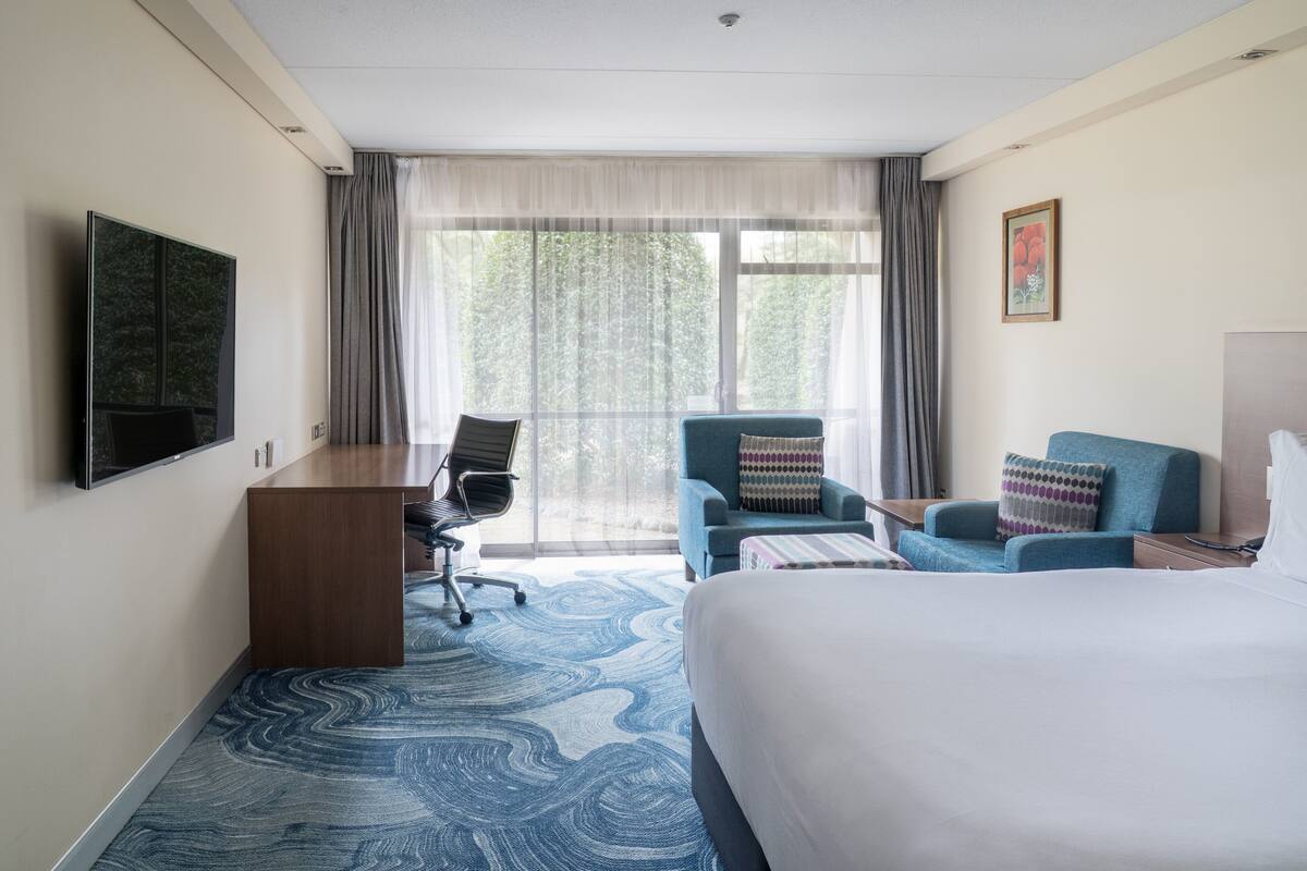 A hotel room with a large bed, a desk, two armchairs, a TV, and a window with curtains.