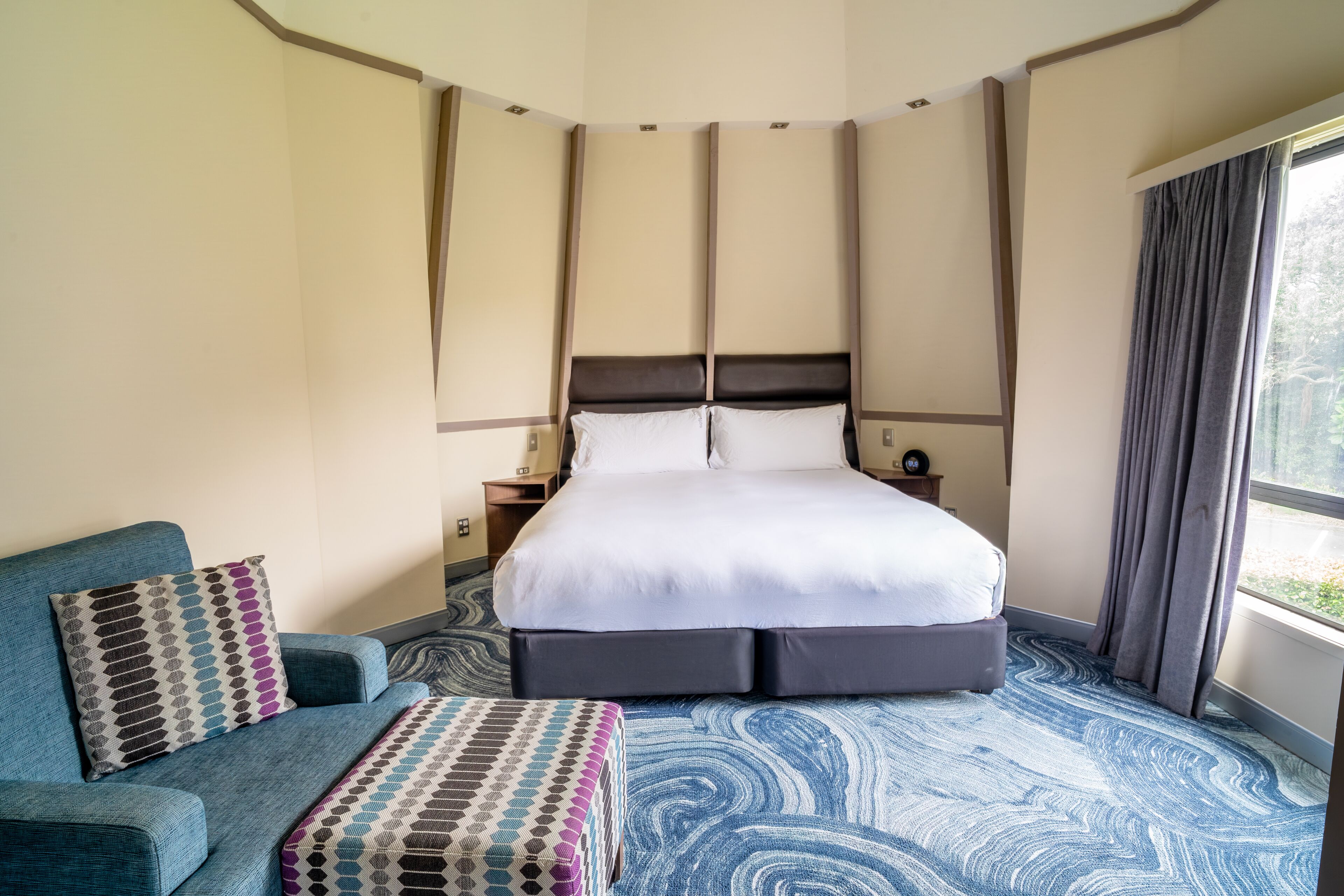 A hotel room with a large bed, a sofa, a patterned ottoman, and a view of greenery through the window.