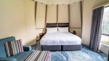 Quarto de hotel com cama grande, sofá, um banquinho estampado e vista para o verde pela janela.